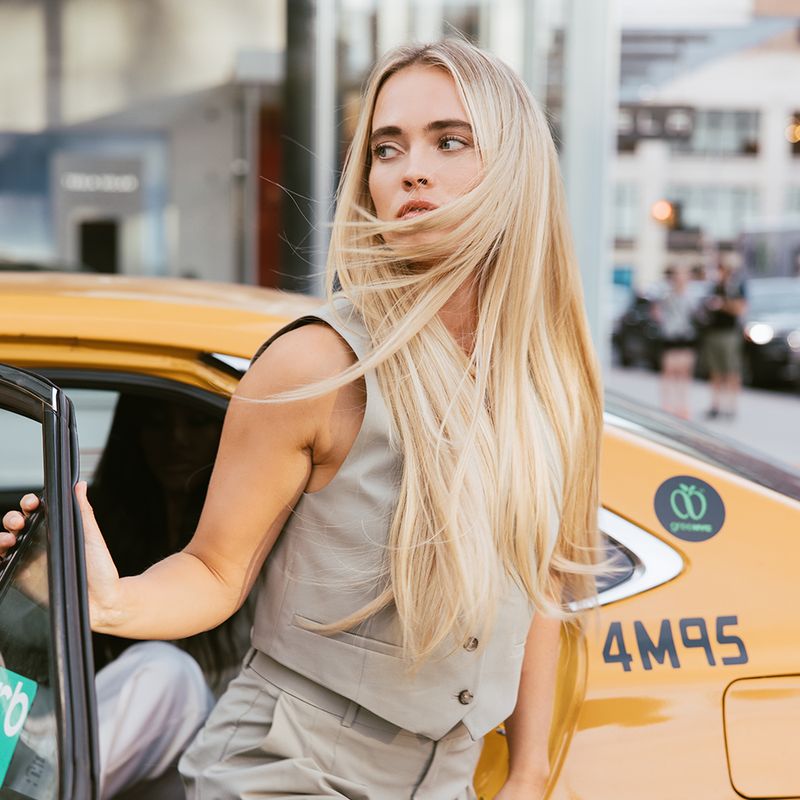 A beautiful woman with long blonde hair extensions from Covet&Mane the Cut Point Silky Straight Collection hair standing next to a taxi cab.